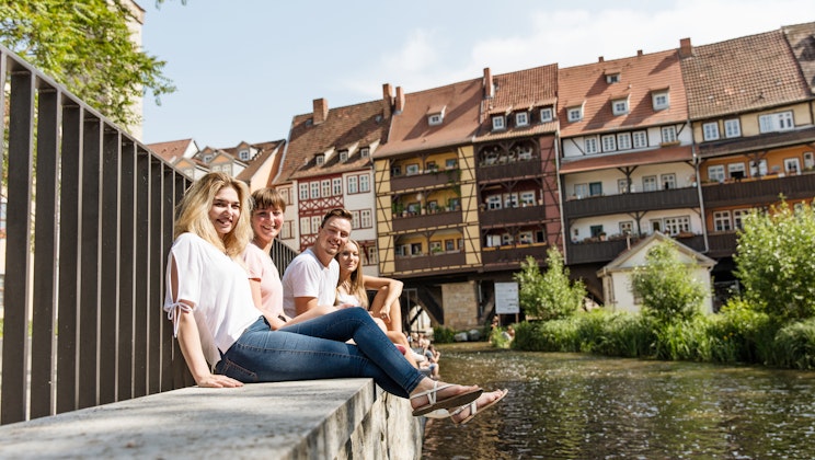 Personen sitzen in Erfurt an einem Fluss, mit der Altstadt im Hintergrund als Symbolbild für das Leben in Erfurt